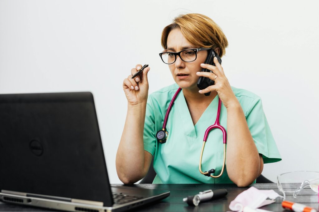 A woman in medical scrubs working with a laptop and phone, illustrating modern healthcare communication.