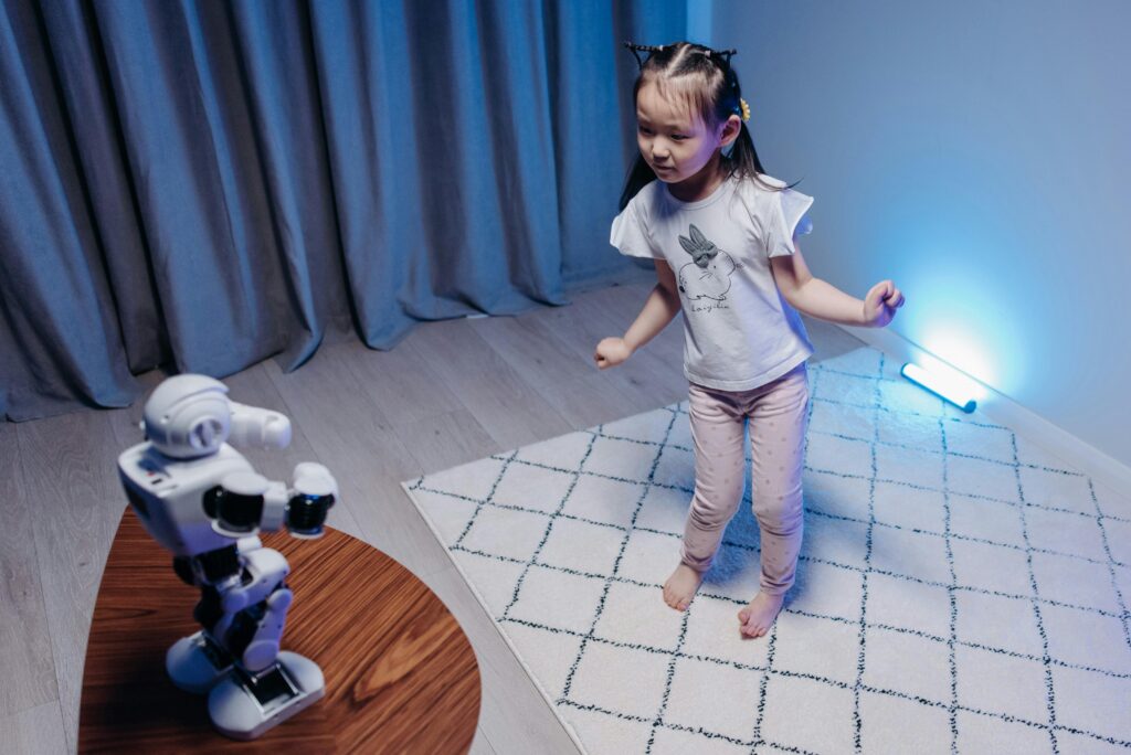 A young girl joyfully dances with a robot on a rug in a modern, softly lit room.